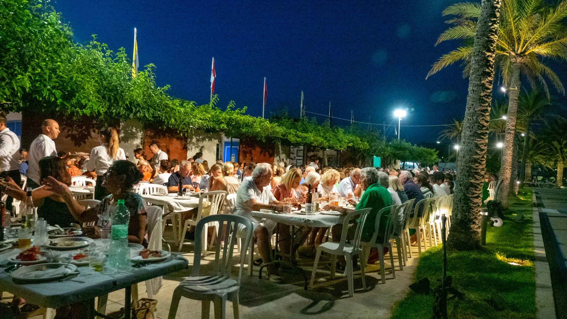 Gente cenando al aire libre durante la noche en un evento social con árboles y palmeras.