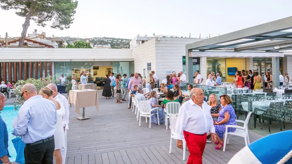 Gente en una terraza al aire libre durante una reunión o evento social, con mesas, sillas y un área de bar, cerca de una piscina, en un día soleado.