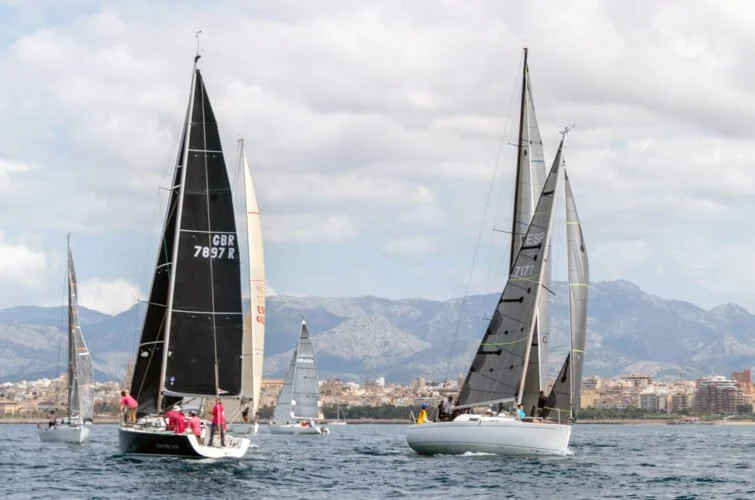 Varias embarcaciones de vela navegando en el mar con una ciudad y montañas en el fondo.