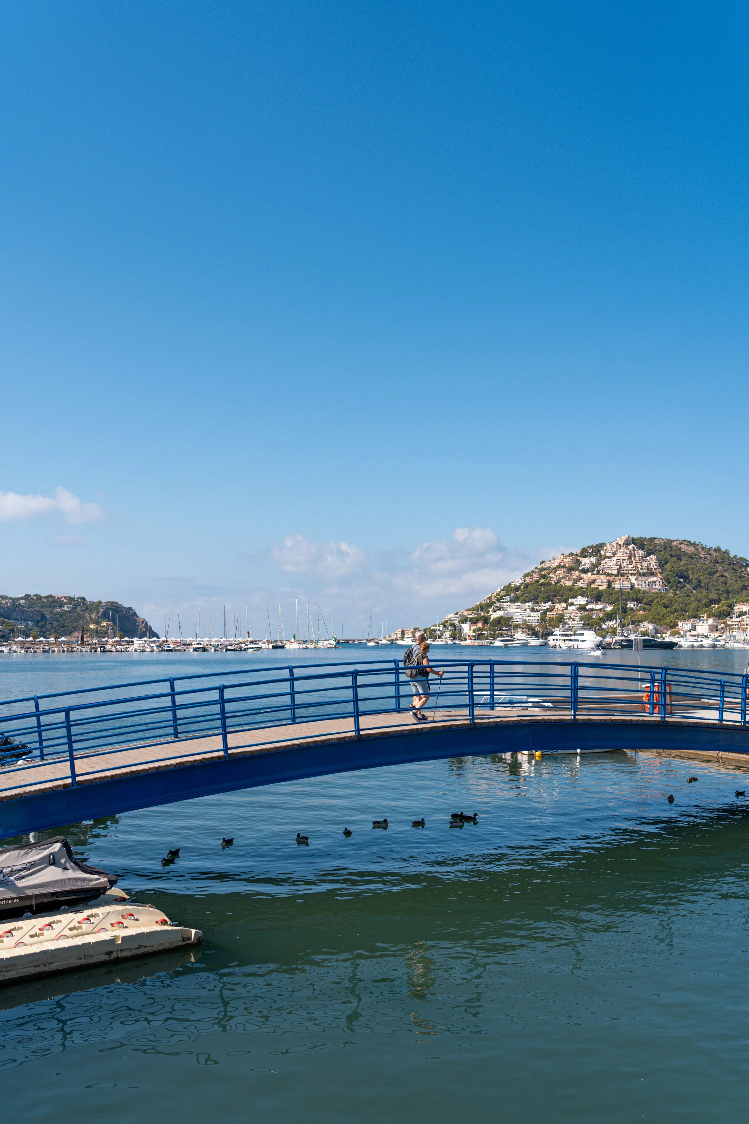 Persona caminando en un muelle de madera con barandillas azules, barcos en el agua y colinas con edificios blancos en el fondo, cielo despejado con pocas nubes.