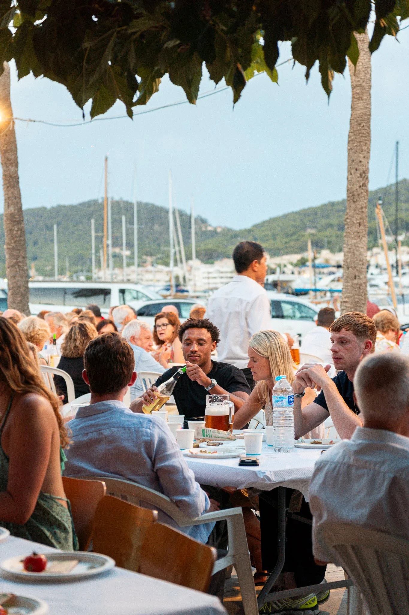 Personas sentadas en una mesa durante una comida al aire libre con vista al puerto con yates y colinas verdes en el fondo, en un ambiente de tarde.