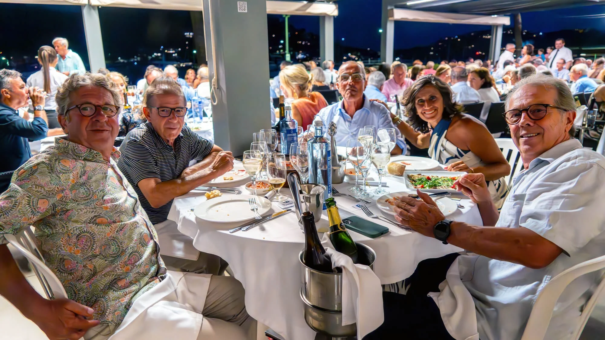 Grupo de personas adultas sentadas alrededor de una mesa en un evento social por la noche, comiendo y bebiendo en un restaurante o terraza abierta.
