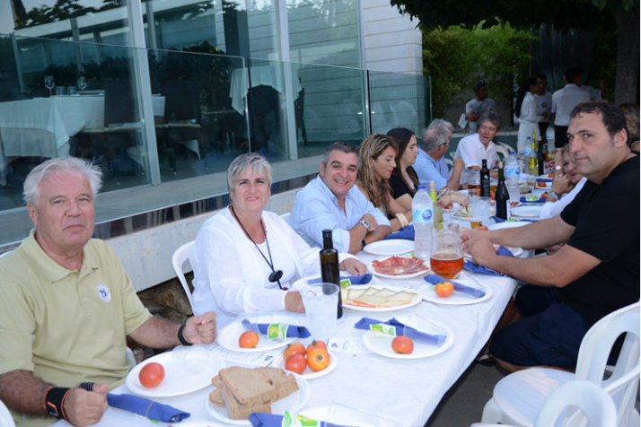 Personas sentadas en una mesa larga disfrutando de comida en un evento al aire libre, con comida, bebidas y decoración.