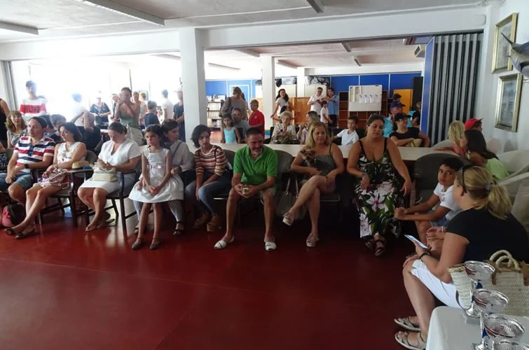 Personas sentadas en sillas en una sala, algunas leyendo, otras usando teléfonos y algunas conversando, con muchas más de pie en el fondo.