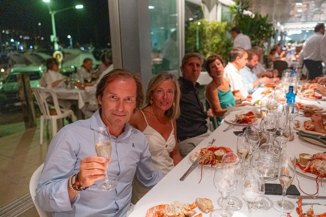 Grupo de personas sentadas alrededor de una mesa con comida y bebidas en un restaurante en la noche.