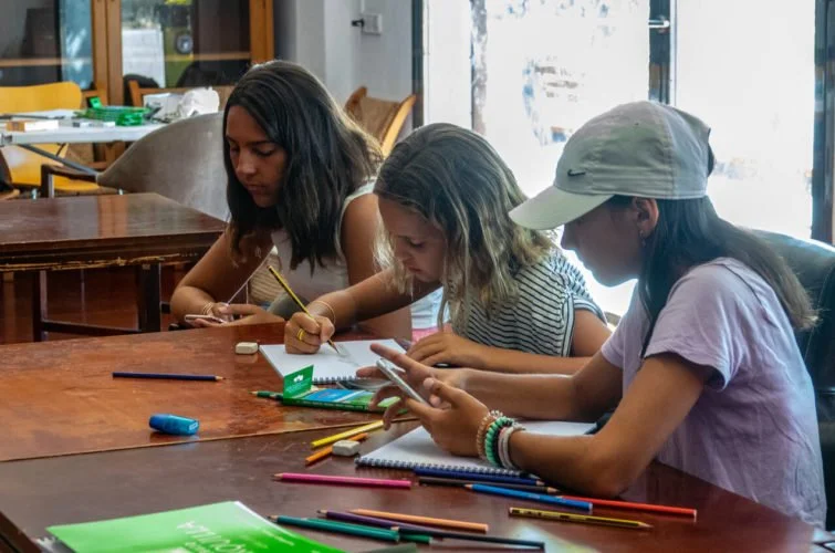Tres niñas en una mesa de madera, una escribiendo, otra con lápiz, y la tercera usando un teléfono móvil, en un ambiente interior con ventanas.