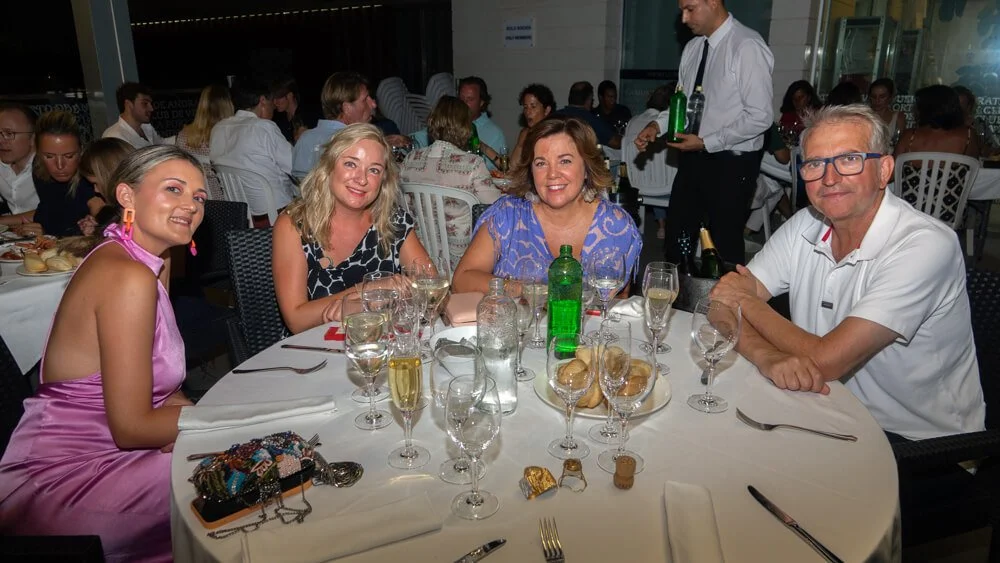 Cuatro personas sentadas en una mesa en un evento social, son dos mujeres jóvenes, una mujer de edad mediana y un hombre, todos sonrientes, con copas y comida en la mesa, en un ambiente de celebración con muchas personas y camareros en el fondo.