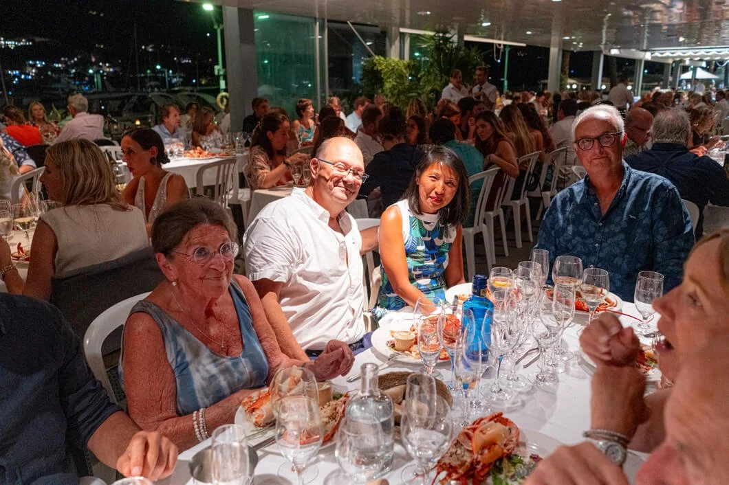 Grupo de personas sentadas en una mesa durante una comida elegante en un restaurante, con muchas copas de vino y platos de comida, fondo con muchas otras mesas y comensales.