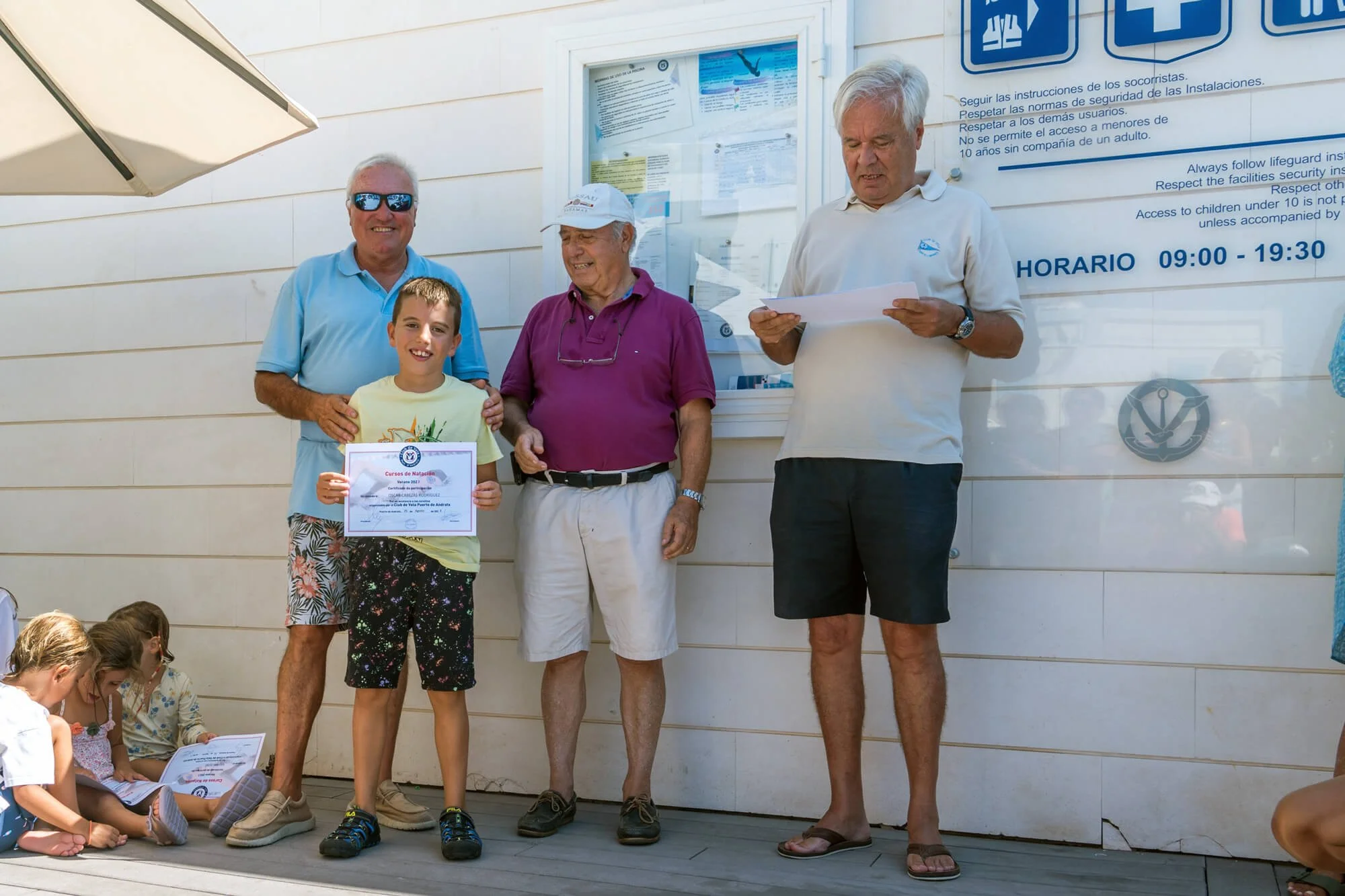 Cuatro personas en un escenario con niños sentados en el suelo, uno de los niños sostiene un certificado, los adultos parecen estar en una ceremonia o premiación en un entorno al aire libre con cartelera de avisos y horario en la pared.