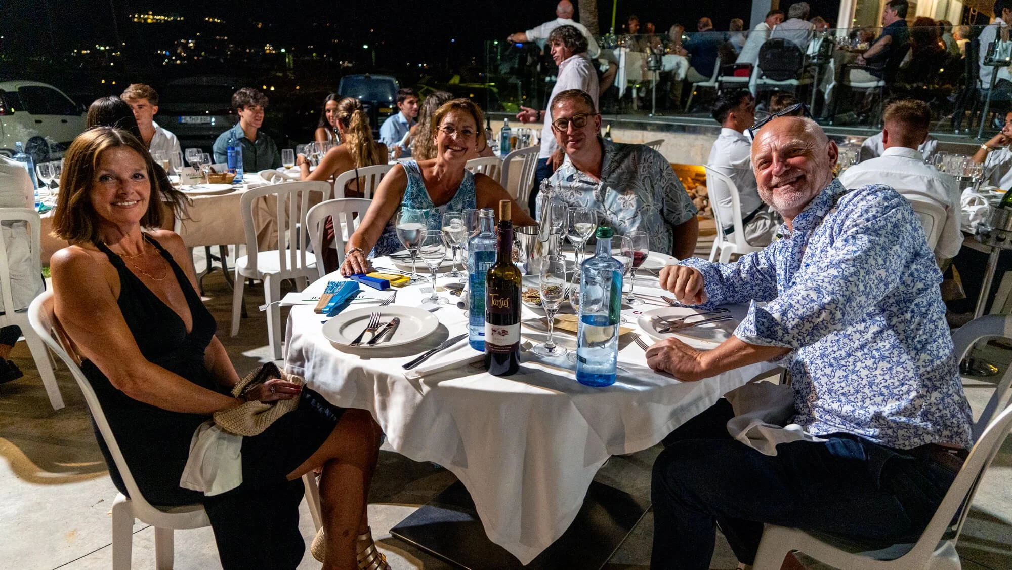 Grupo de personas sentadas en una mesa durante una cena en un restaurante al aire libre en la noche, con fondo de luces y otros comensales.