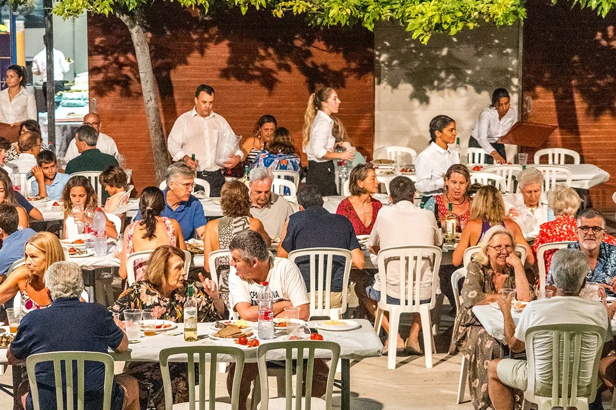 Una reunión al aire libre con muchas personas sentadas en mesas comiendo y bebiendo, con algunos meseros atendiendo y un ambiente social animado.