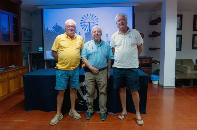 Tres hombres (uno vestido con camiseta amarilla y pantalones cortos, otro con camisa azul y pantalones beige, y el tercero con camiseta blanca y pantalones cortos) posando en un interior con pantalla y suelo de madera.