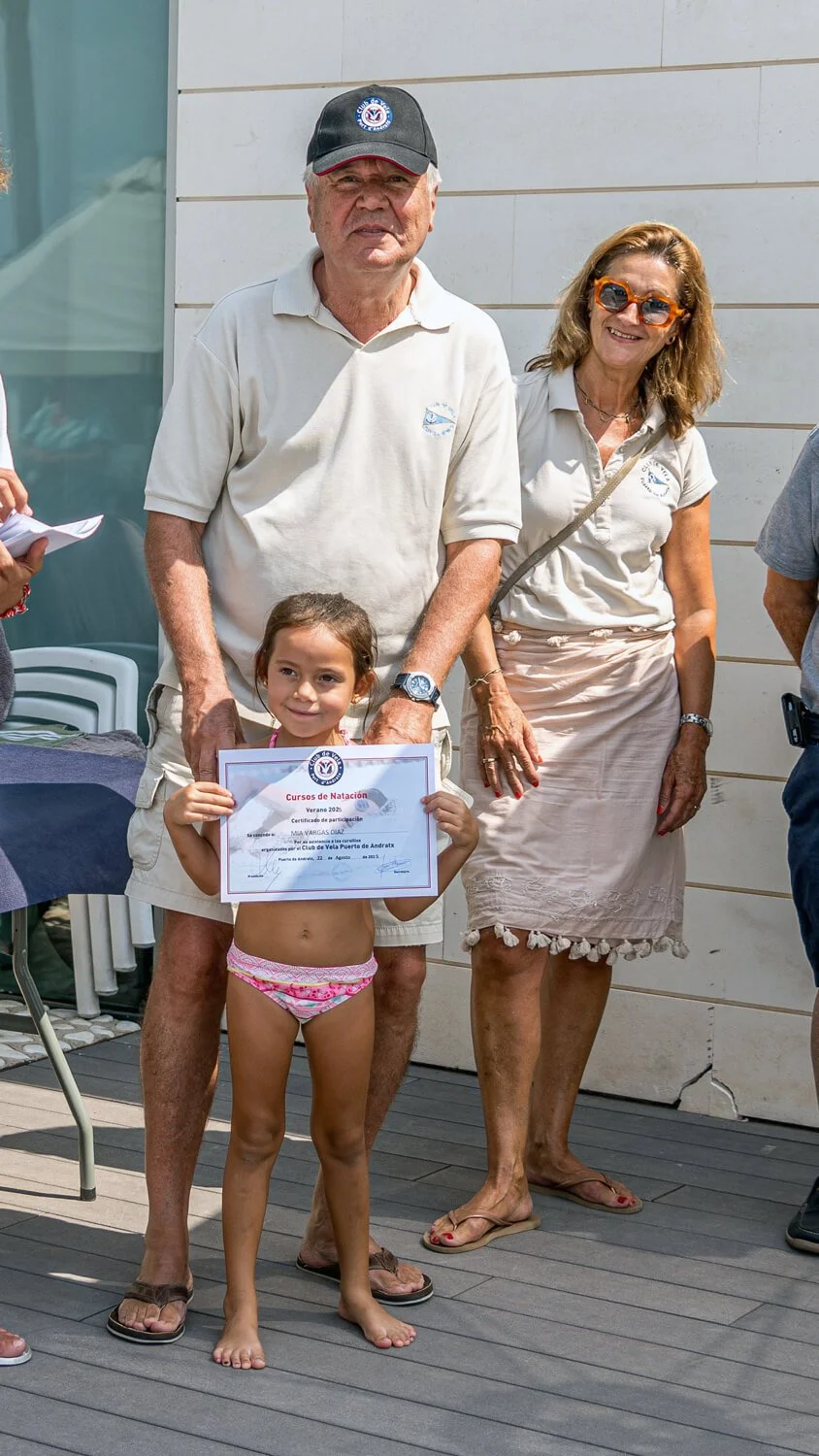 Una niña con traje de baño recibe un certificado de participación en un evento de natación, acompañada por un hombre mayor y una mujer, en un ambiente de celebración al aire libre.