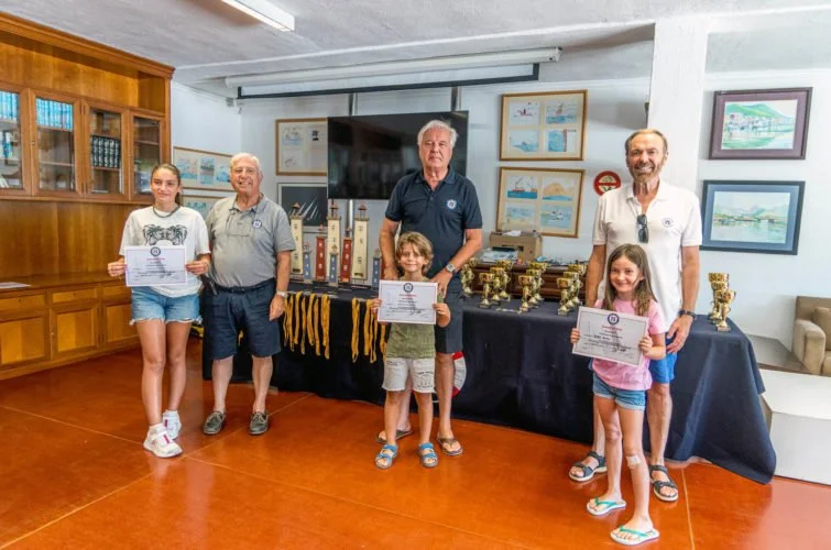 Tres niños y tres adultos en una ceremonia de entrega de premios en un salón con muebles de madera y cuadros en la pared, sosteniendo diplomas y trofeos en una mesa con más trofeos.