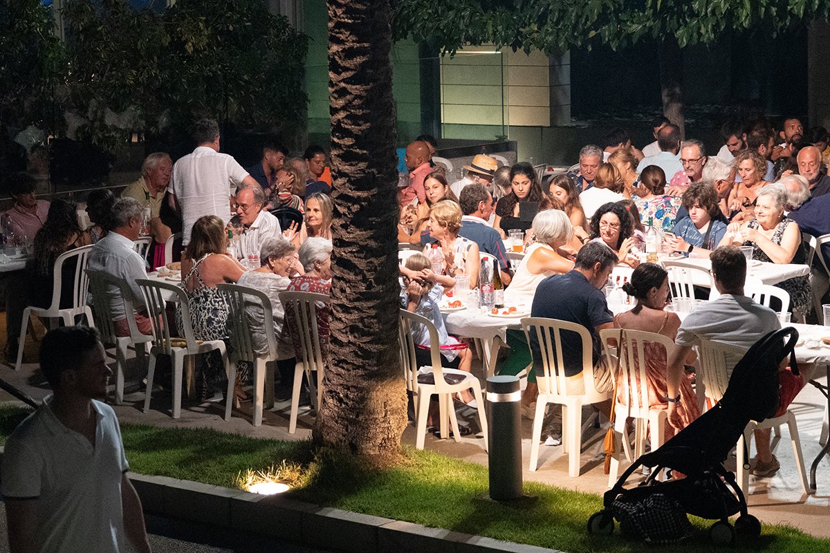 Reunión social al aire libre en la noche con muchas personas sentadas en mesas disfrutando comida y bebida, en un entorno con palmeras y césped.