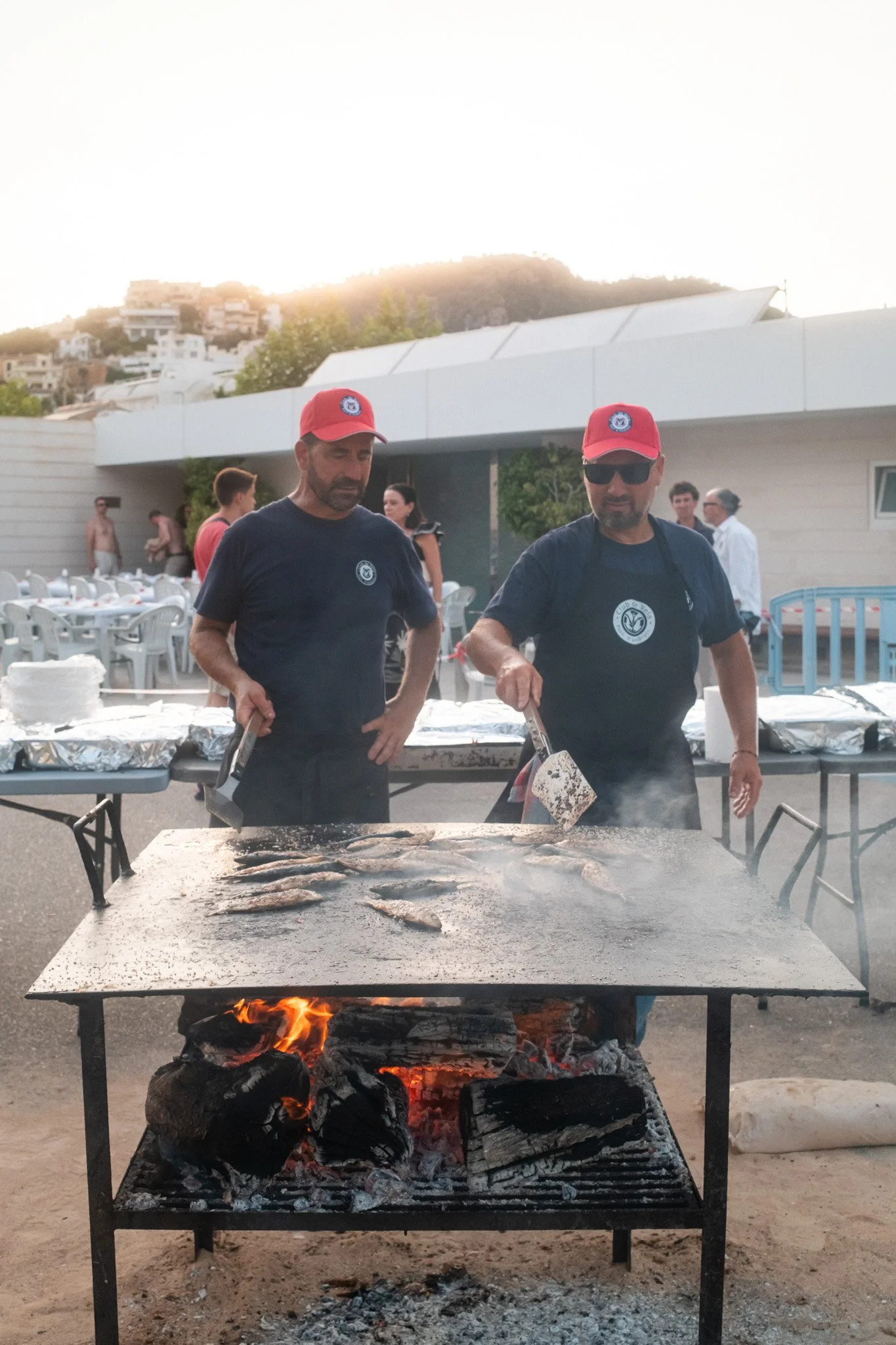 Dos hombres cocineros preparando comida en una parrilla gigante con carbón y fuego, en un evento al aire libre, en un entorno con montañas y casa blanca en el fondo, durante el atardecer.