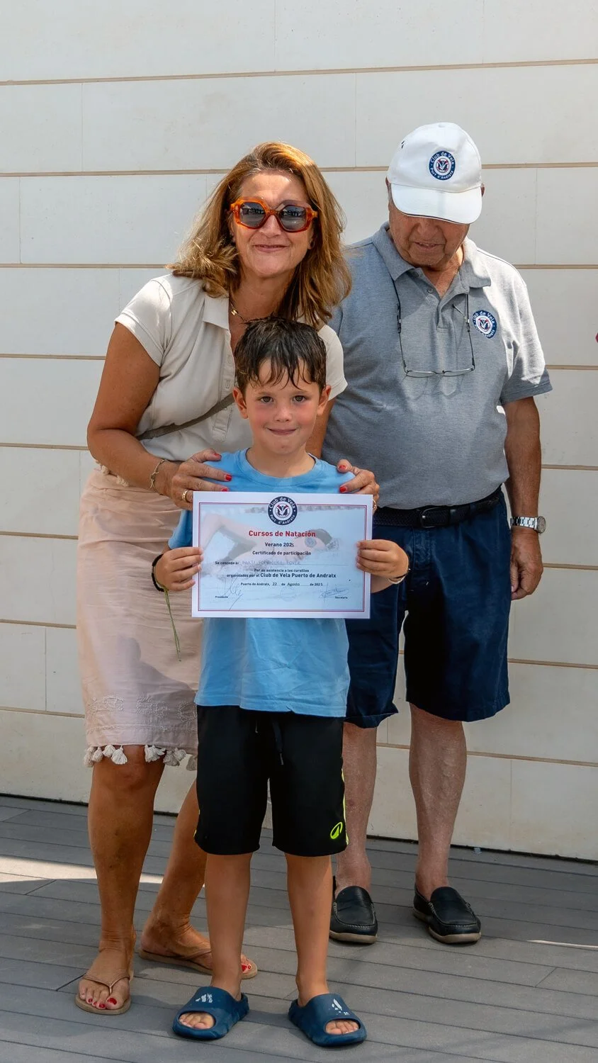 Un niño sosteniendo un certificado de participación en cursos de natación, flanqueado por dos adultos en un evento de natación, todos sonrientes, con un fondo de pared blanca.