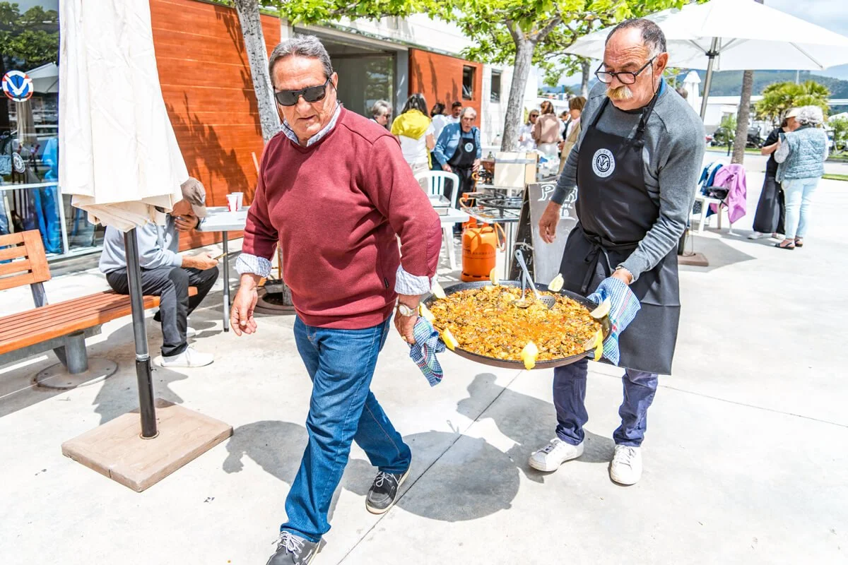 Hombre con gafas de sol y suéter rojo lleva un plato grande de comida, mientras que un hombre con gafas y delantal negro le ayuda en una terraza al aire libre.