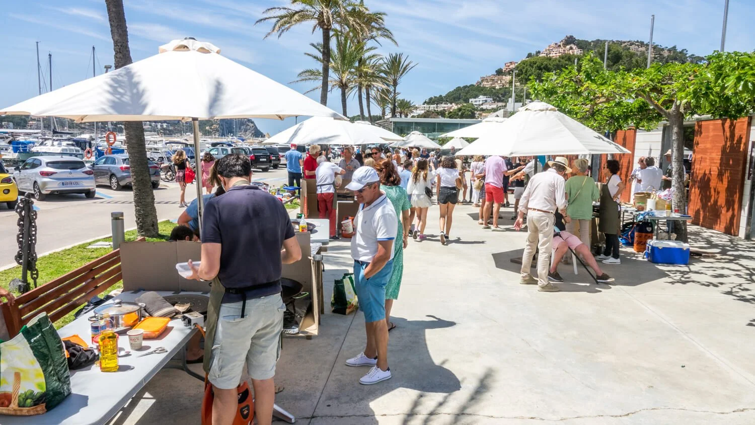 Personas en mercado al aire libre con sombrillas, en un puerto con yates y colinas con casas en el fondo, en un día soleado y despejado.