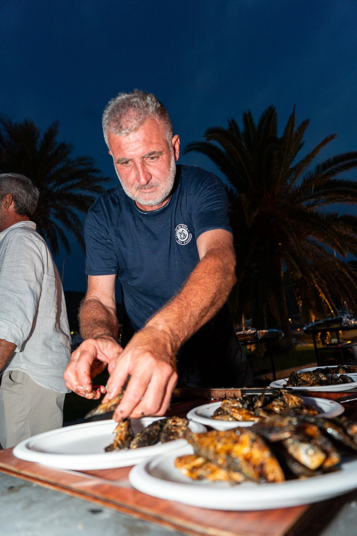 Hombre sirviendo y colocando mariscos en platos en una fiesta al aire libre durante la noche, con palmeras en el fondo.