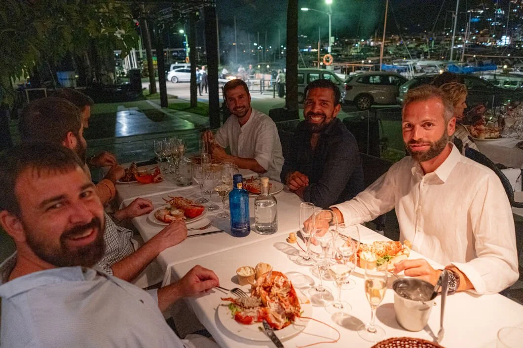 Un grupo de personas cenando en un restaurante al aire libre por la noche, con comida y bebidas en la mesa y un fondo de barcos en un puerto.