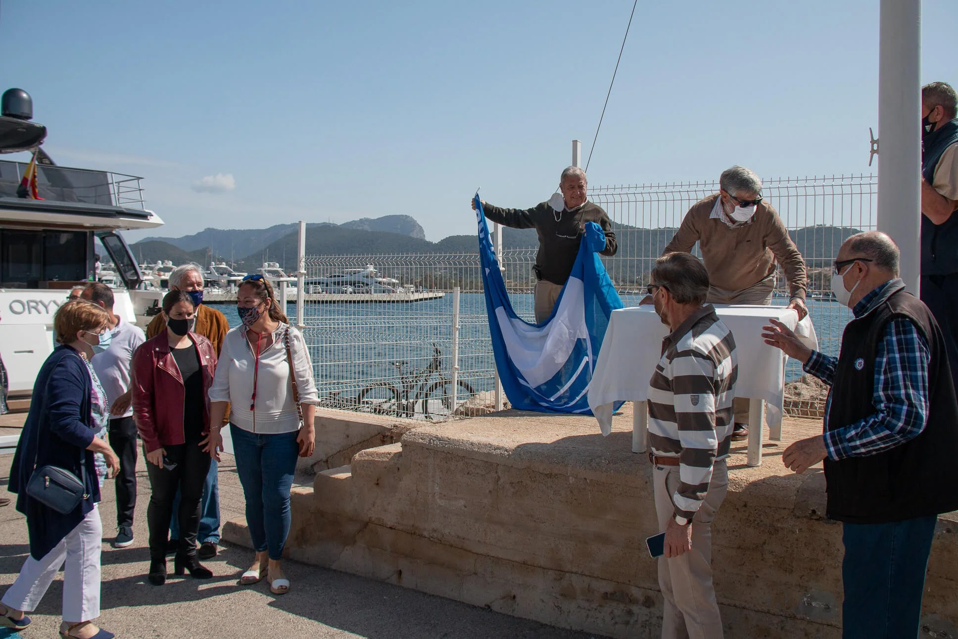 Grupo de personas en una ceremonia al aire libre junto a un puerto, con fondo de montañas y barcos. Algunas personas llevan mascarillas y están alrededor de una mesa cubierta con una bandera y un objeto decorativo.
