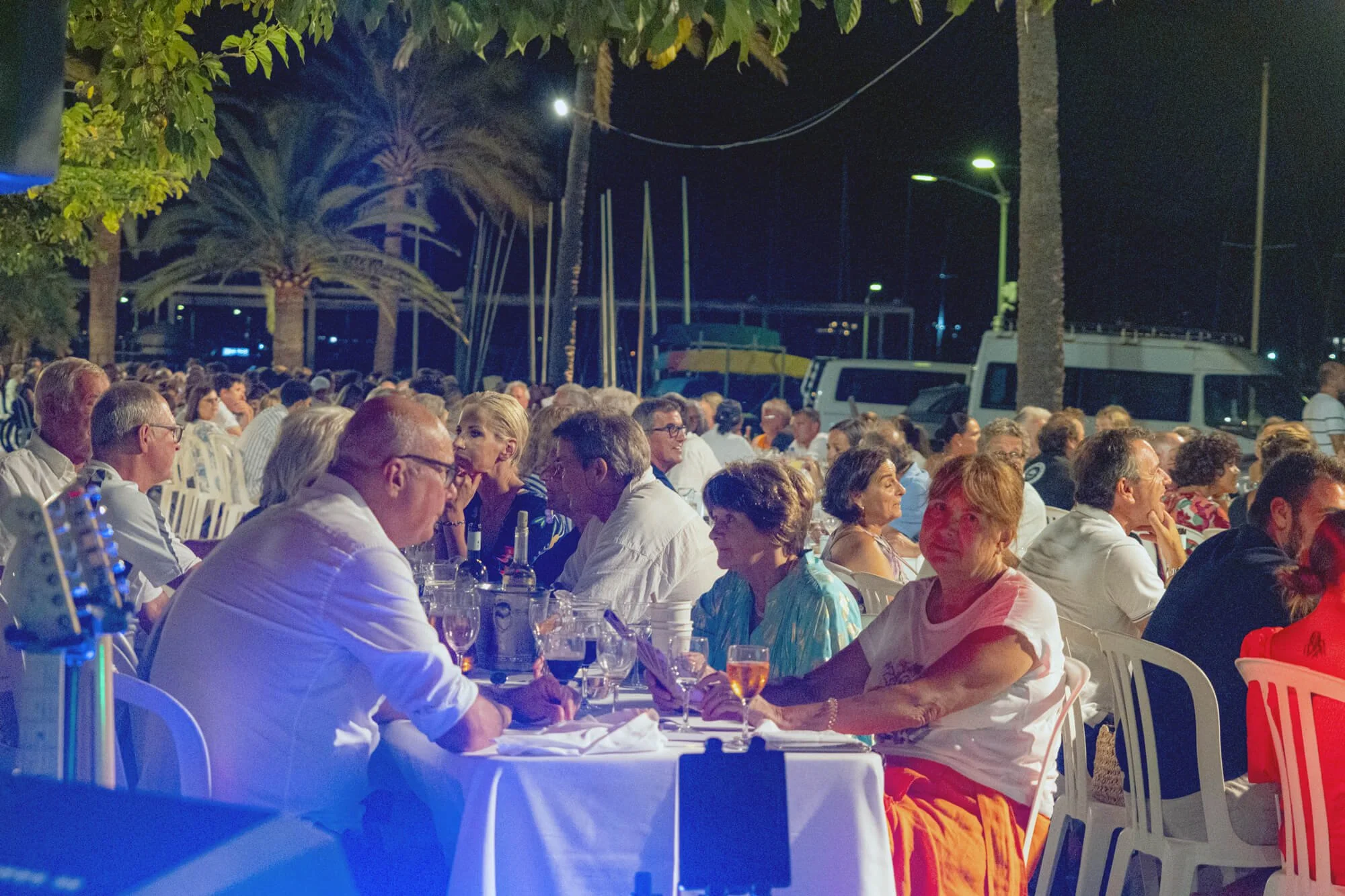 Personas sentadas en una mesa durante una reunión nocturna al aire libre, con árboles y barcos en el fondo, iluminadas por luz artificial.