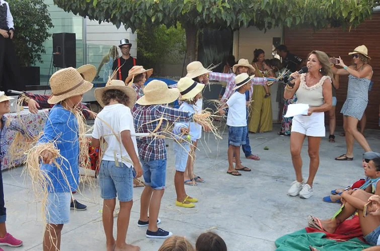 Niños en un festival con sombreros y ropa de campo, participando en actividades tradicionales, con una mujer que habla con micrófono y un grupo de personas en el fondo.