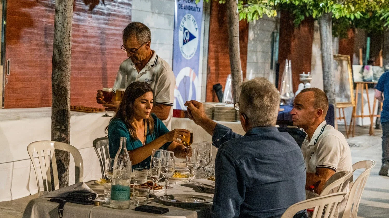 Una escena en una cena al aire libre, con cuatro personas alrededor de una mesa y un camarero sirviendo bebidas en el fondo. La mesa tiene varios vasos, botellas y comida.