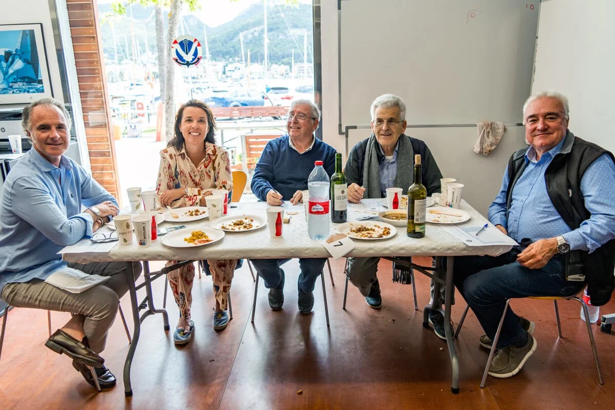 Cinco personas sentadas en una mesa de comedor con comida y bebidas, en un interior con ventana que muestra un paisaje con montañas y barcos.