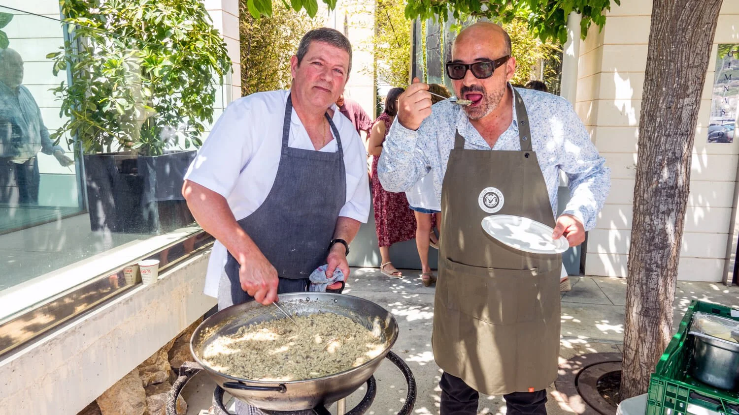 Dos hombres cocinando y disfrutando al aire libre, uno con una sartén grande y el otro comiendo con una cuchara, ambos usando delantales. Hay árboles y personas en el fondo.