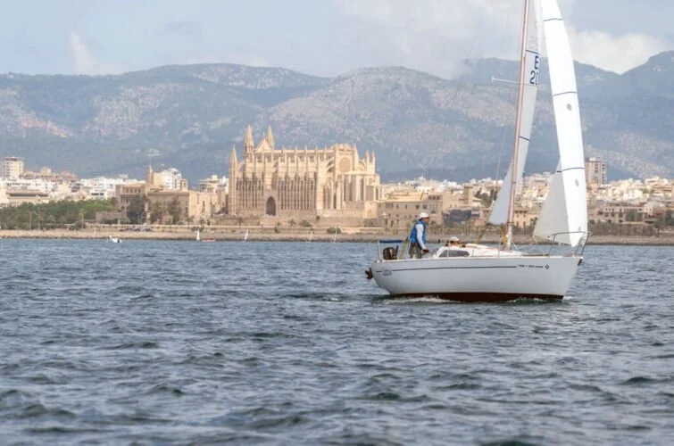 Velero en el agua con ciudad y catedral en el fondo, con montañas al fondo en clima nublado.