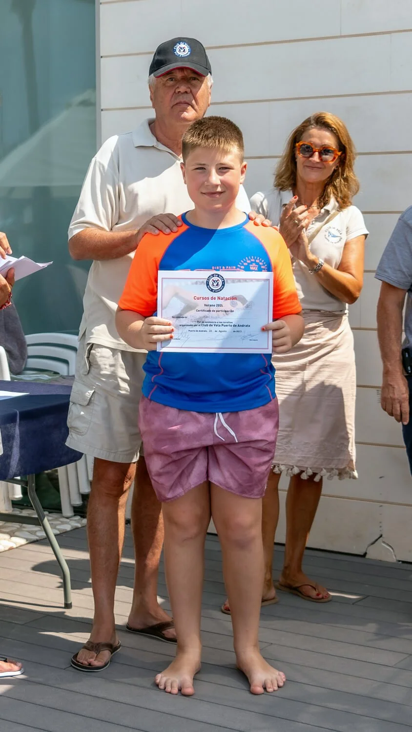 Un niño sosteniendo un certificado de participación en cursos de natación, acompañado por dos adultos en un evento al aire libre con paredes blancas y suelo de madera.