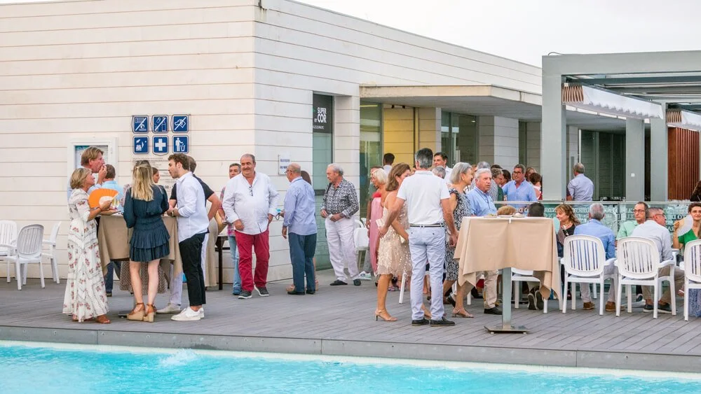 Personas socializando en un evento junto a la piscina, con mesas y sillas, en un espacio exterior y moderno.