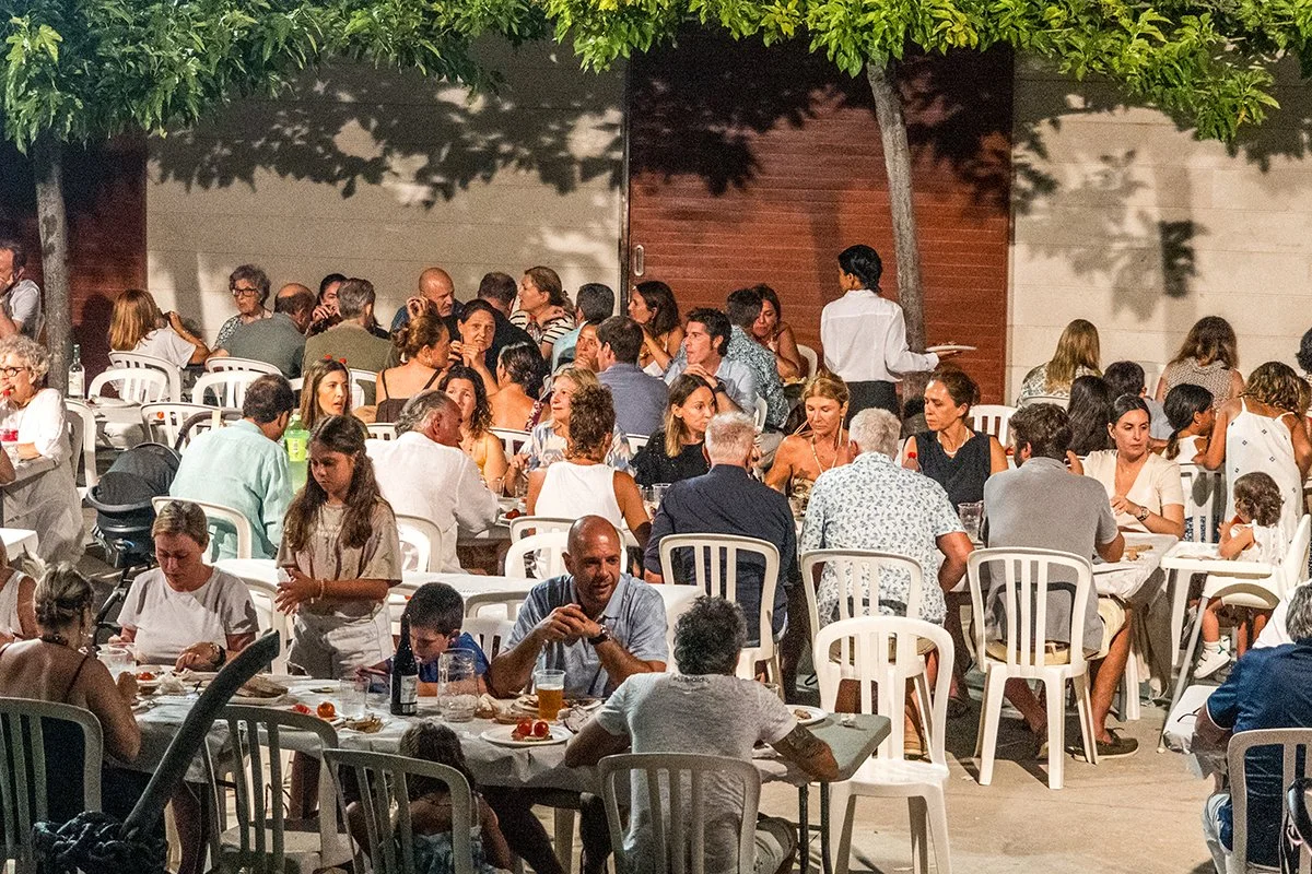 Gente disfrutando en una comida al aire libre en la noche, con muchas mesas y sillas, árboles y paredes de fondo.