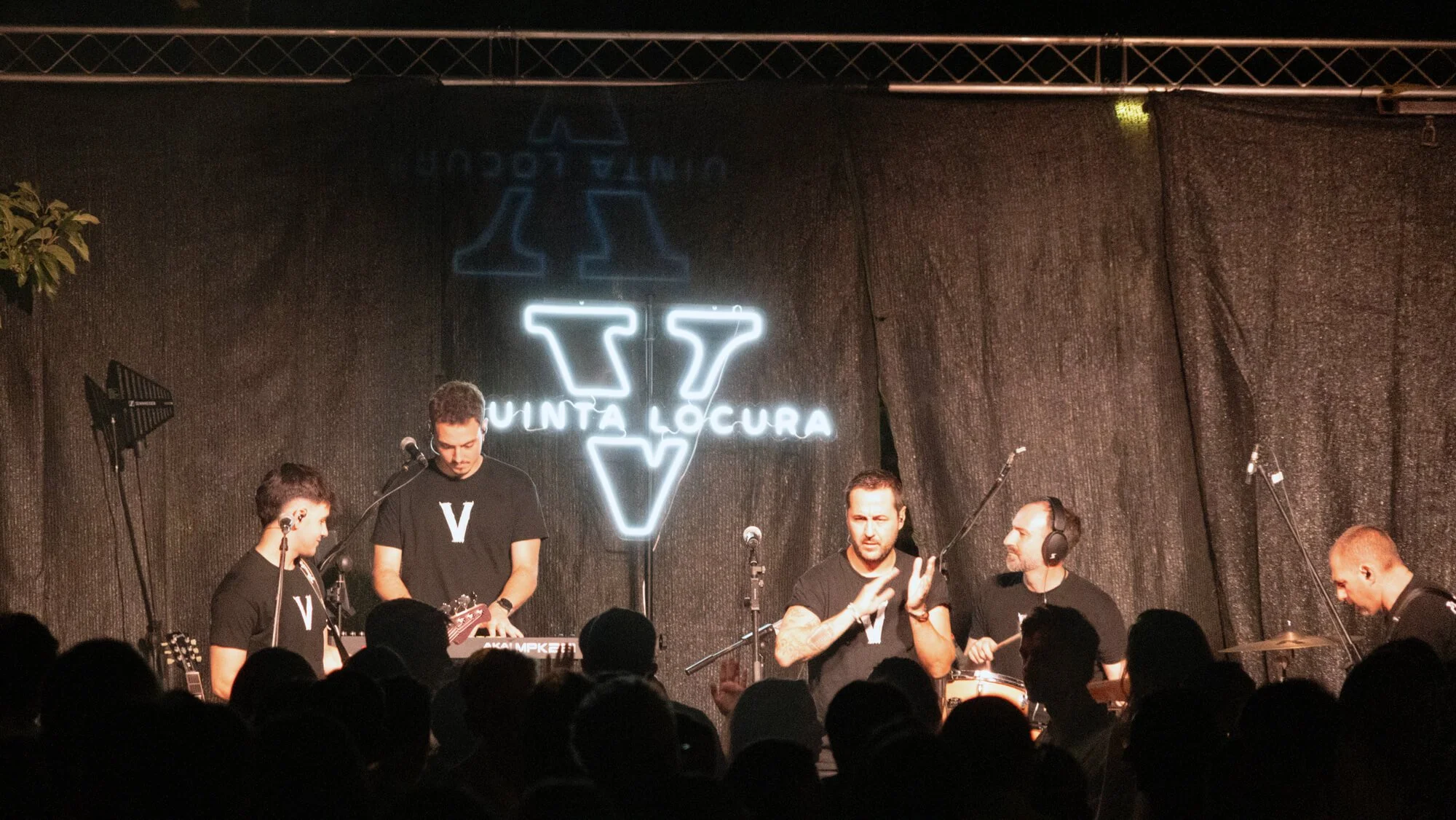 Grupo de personas tocando instrumentos en un escenario con fondo negro y cartel de neón que dice 'Vunta Locura'.