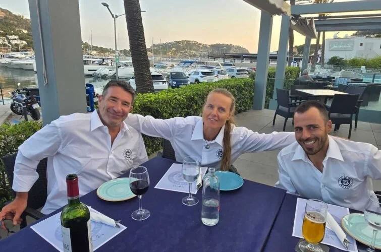 Tres personas sentadas en una mesa de restaurante frente a un puerto con barcos y colinas al atardecer.