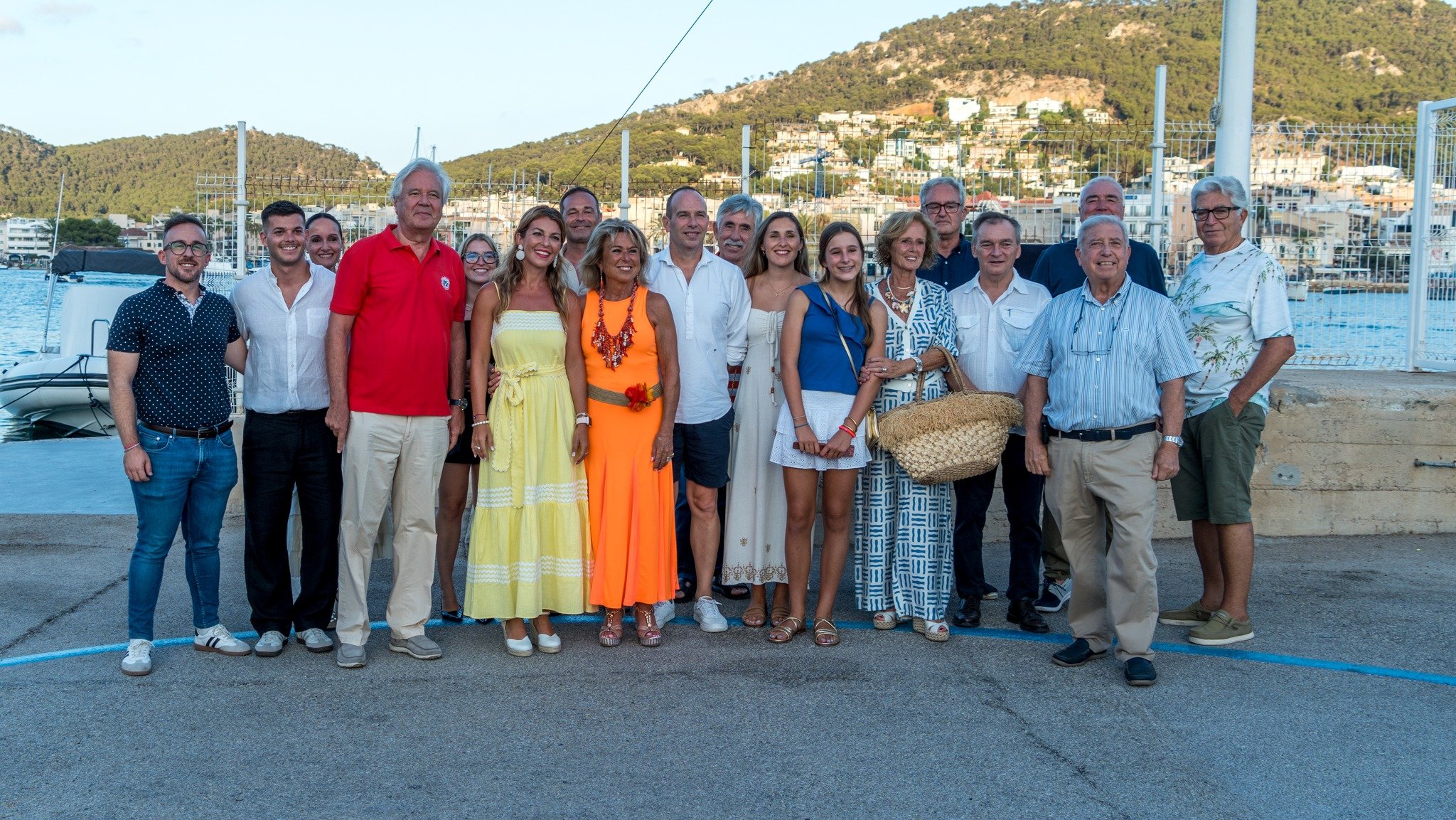 Grupo de personas posando en un puerto con montañas al fondo, algunos vistiendo ropa colorida y otros en tonos neutros, con barcos y casas en la terraza de las colinas.