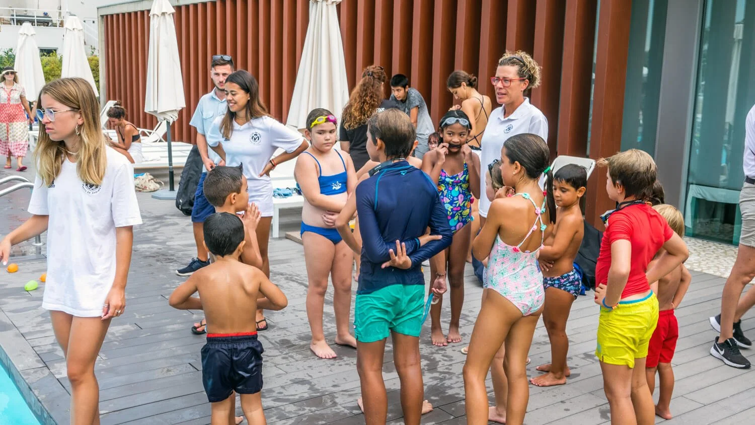 Niños en ropa de baño y adultos en una reunión junto a la piscina cubierta de un área de descanso con paredes de madera y sombrillas cerradas.