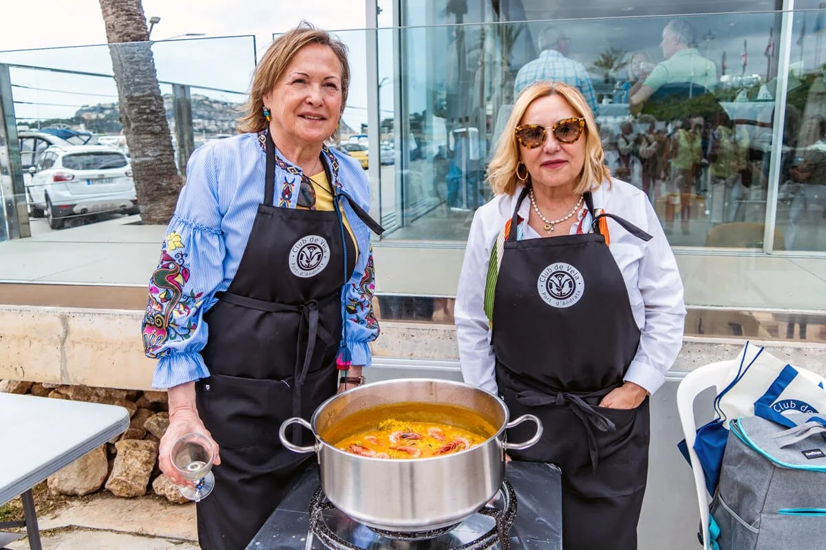 Dos mujeres mayores con delantales negros preparando una sopa en una olla grande al aire libre, frente a un edificio de cristales con muchas personas y autos en el fondo.