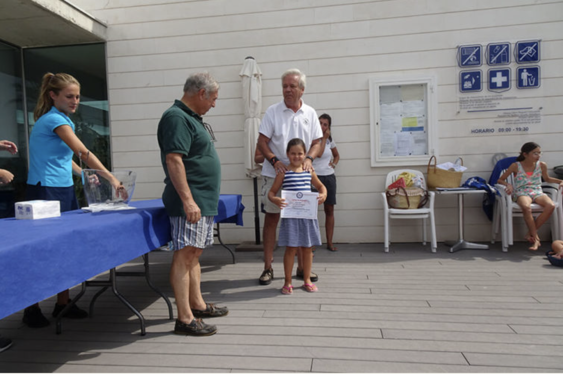Una ceremonia de premiación infantil en un espacio al aire libre con varias personas, niños y adultos. Una niña con vestido a rayas recibe un reconocimiento en manos de un adulto. Hay una mesa con un mantel azul a la izquierda, y en el fondo un panel