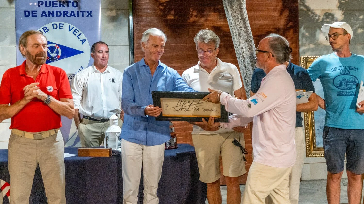 Grupo de hombres en una ceremonia, algunos sosteniendo un cuadro y otros observando, en un entorno con logotipo de club de vela y decoración de madera.