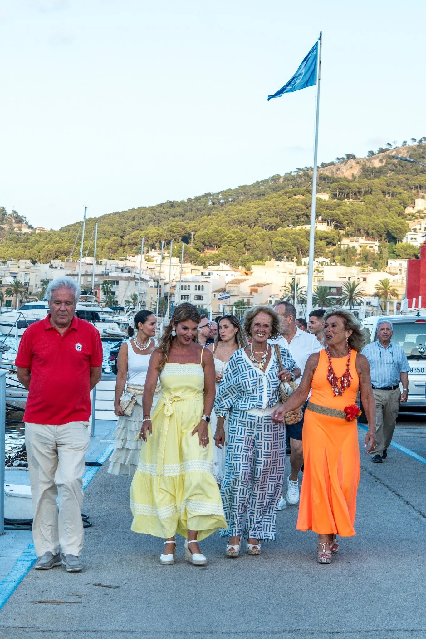 Grupo de personas caminando cerca de un puerto con yates y montañas verdes en el fondo, todas vestidas de colores claros y alegres en un día soleado.