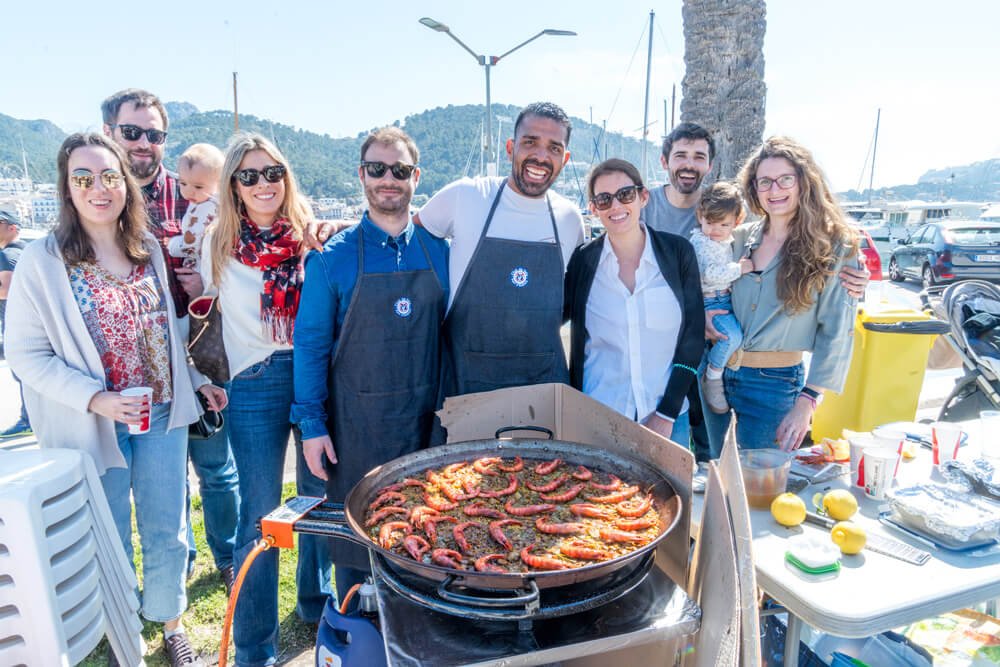 Grupo de personas sonriendo en una reunión al aire libre con un chef cocinando mariscos en una plancha. Están cerca del mar, en un puerto con barcos, y hay una mesa con comida y utensilios.