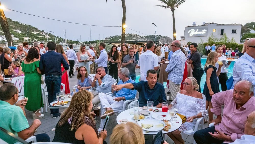 Personas en una reunión social al aire libre con mesas y bebidas, en un entorno junto a un cuerpo de agua y palmeras.