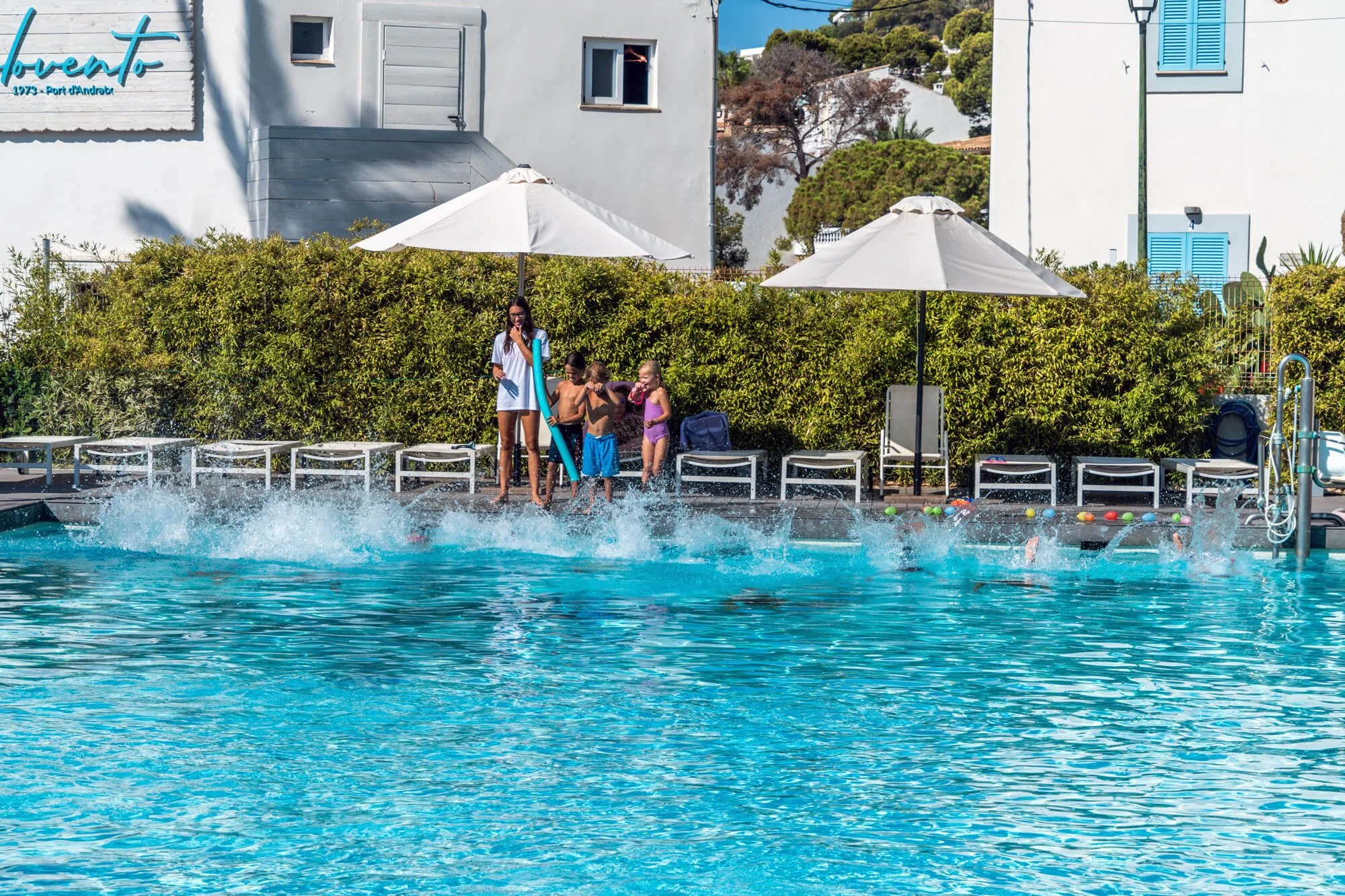 Niños jugando en una piscina con paraguas blancos, frente a un seto verde y casas blancas con ventanas azules.