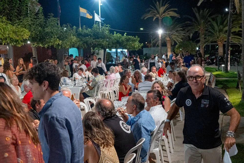 Multitud de personas sentadas y de pie en una fiesta al aire libre durante la noche, con muchas mesas con comida y bebida, palmeras y banderas en el fondo.
