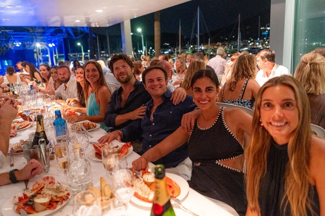 Grupo de personas disfrutando de una cena en un restaurante con vista a un puerto en la noche, con mariscos y bebidas en la mesa.
