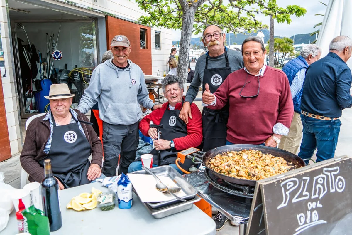 Grupo de cinco personas sonriendo en una reunión al aire libre, con un cartel que dice "Pizza del día" y una paella gigante en una mesa.