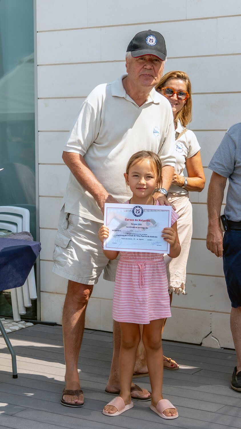 Una niña recibe un certificado de participación en un curso de natación, acompañado por un hombre mayor y varias personas detrás, en un evento al aire libre.